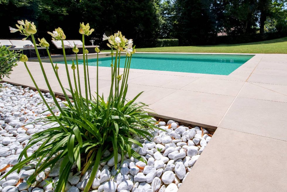 Détail de la plage de piscine en grès cérame et du pourtour paysagé avec galets blancs et plantes, projet Mondial Piscine à Buellas