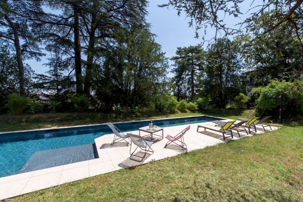 Terrasse aménagée avec transats au bord de la piscine couloir de nage à Beynost, dans un jardin arboré